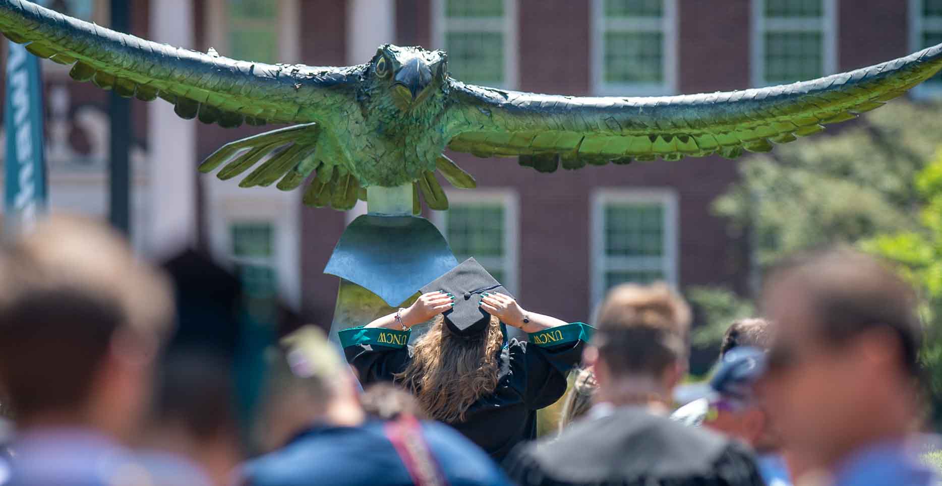 Student holds graduation cap and looks up at UNCW seahawk statue, as crowd from graduation ceremony walks by her.