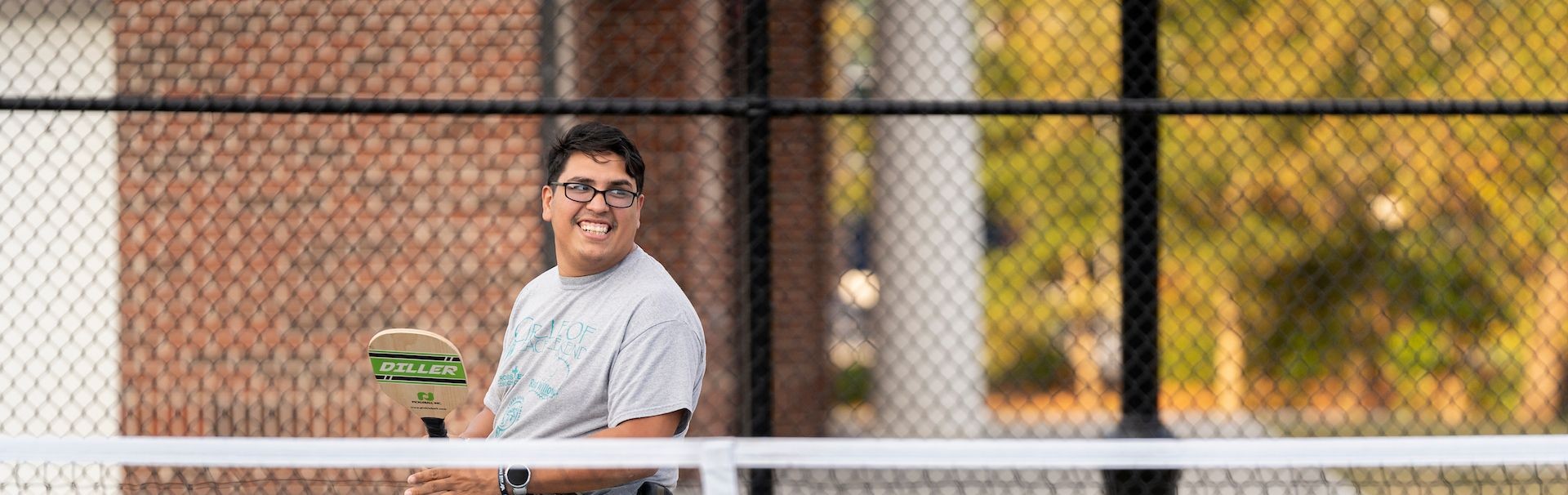 A smiling wheelchair user holds a pickleball paddle on an outdoor court.