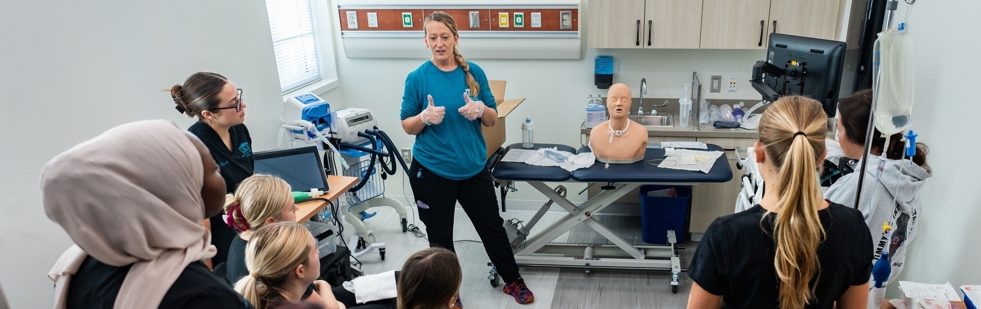 Respiratory students observe to their professor teach about tracheostomy on a manikin.