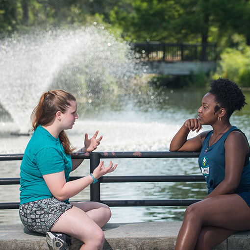 Two students talking while sitting on the edge of a water fountain.