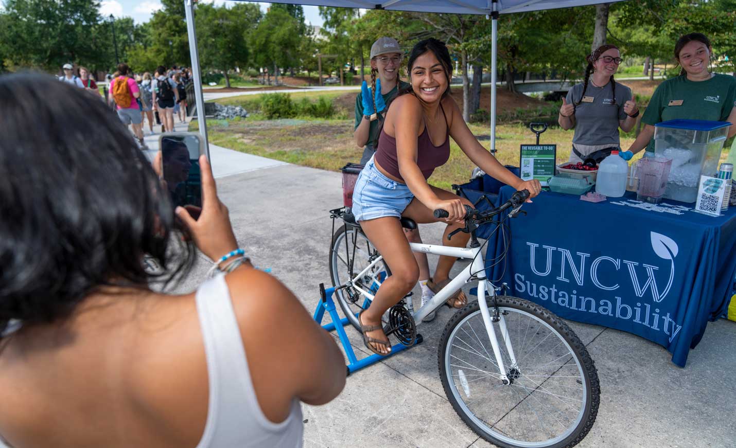 A female presenting person rides a bike with a blender on the back in front of an outreach table. 