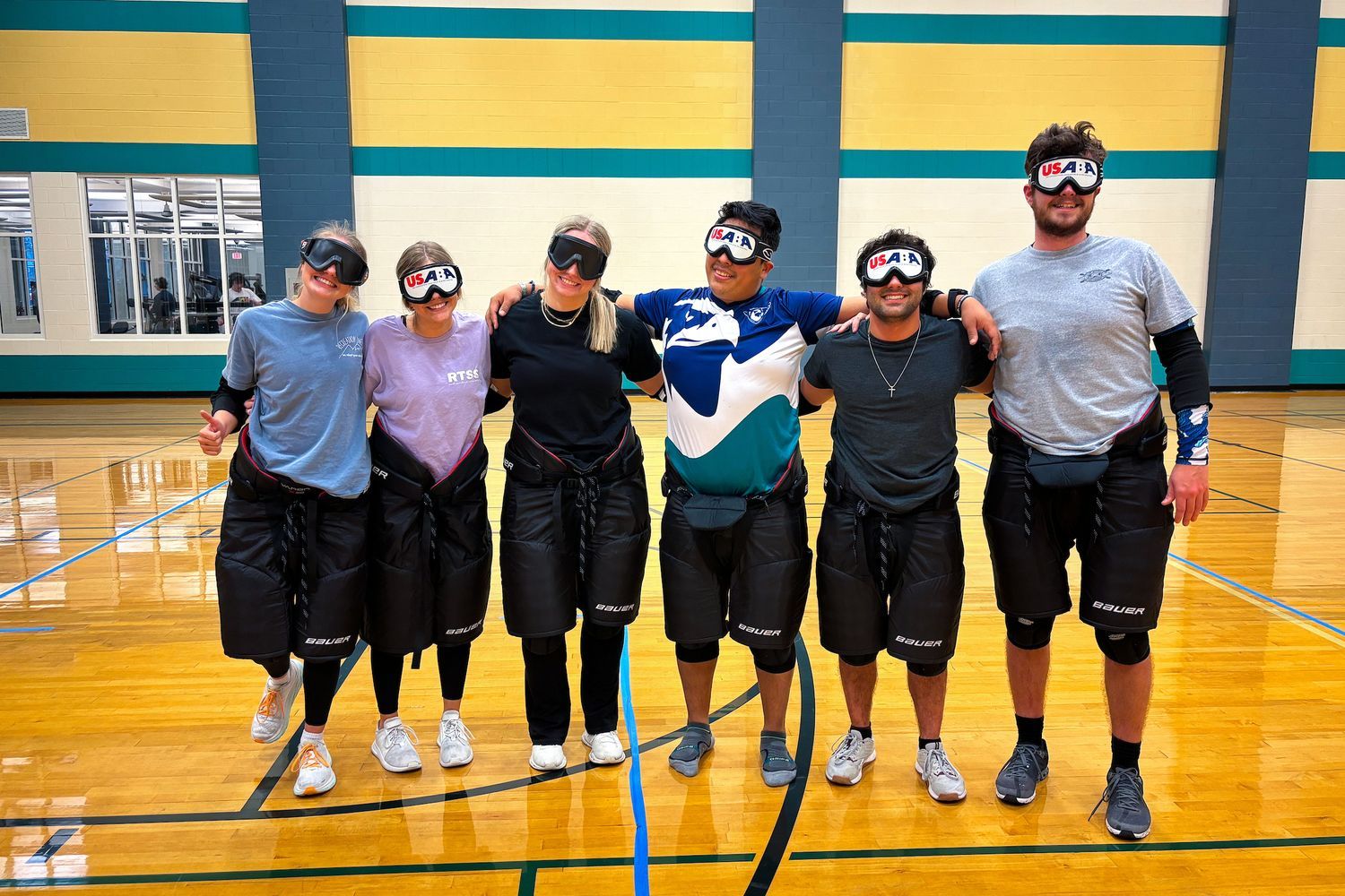 six students pose in the gym while wearing goal ball gear, which includes goggles they cannot see out of, elbow pads and knee pads.