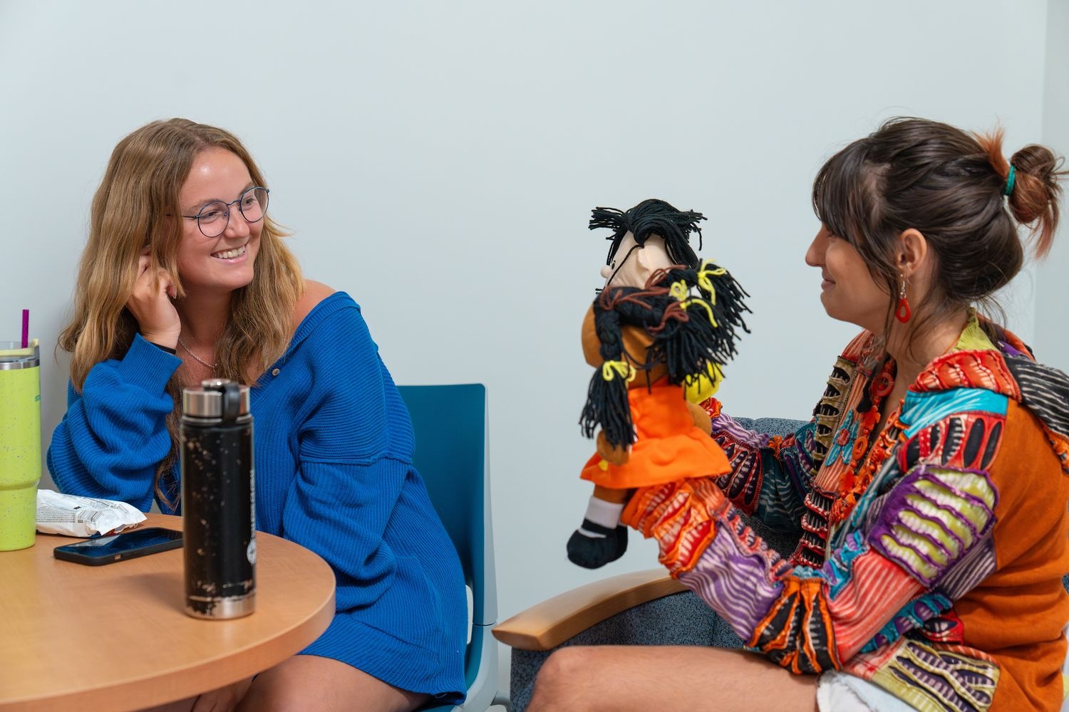 two master of social work students practice scenarios together. One student holds up two puppets while the other student smiles.