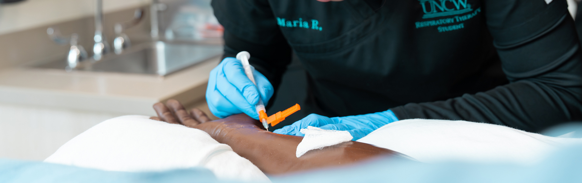 the gloved hands of a respiratory therapy student practices drawing blood from the radial artery of a manakin. 