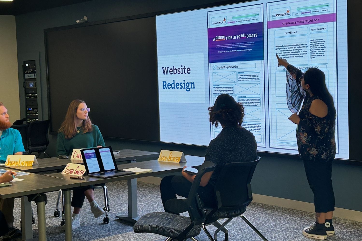 Person presenting a website redesign mockup on a large screen during a meeting while attendees watch from a conference table.