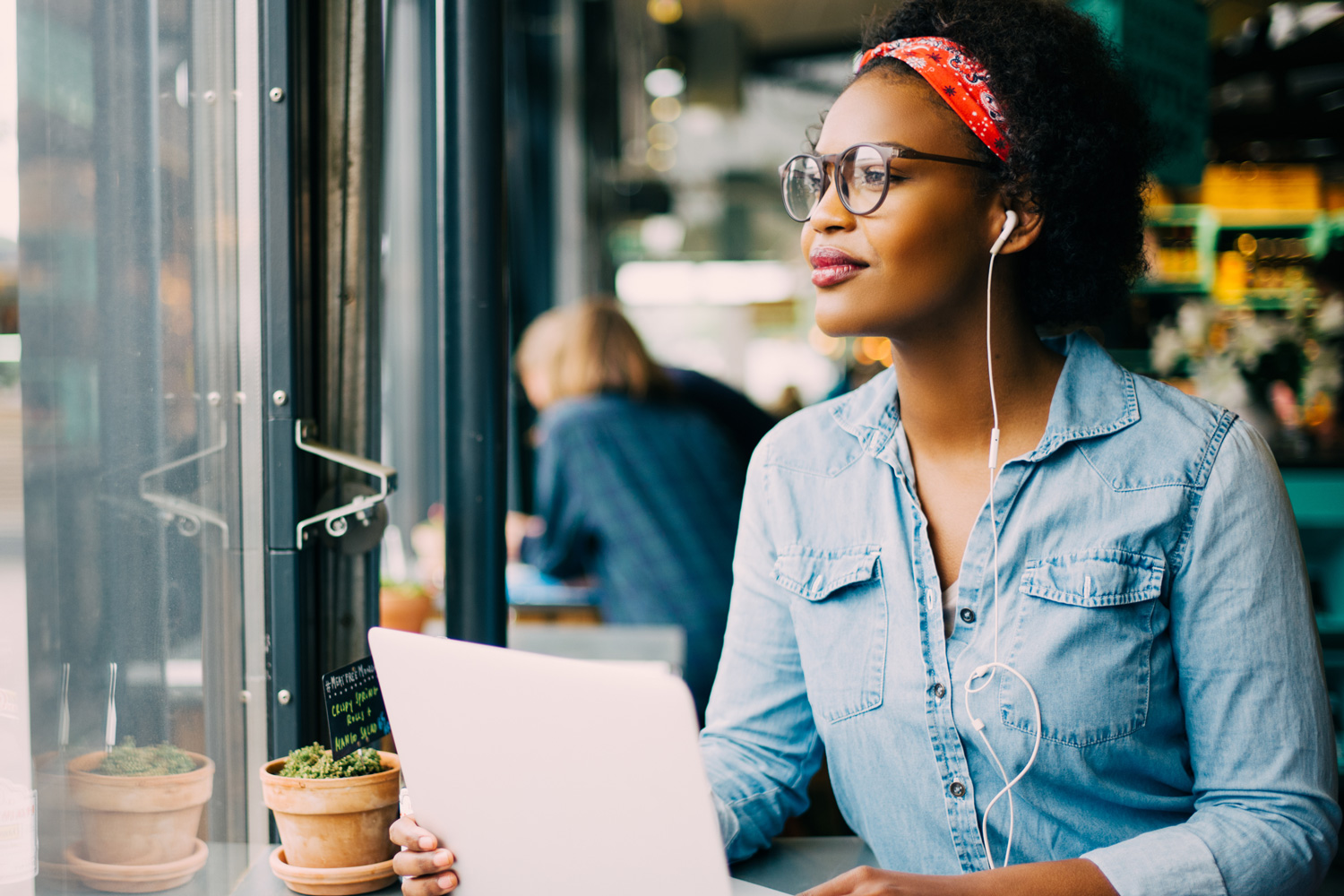 Smiling young African woman looking through a window while sitting alone in cafe working on a laptop and wearing headphones