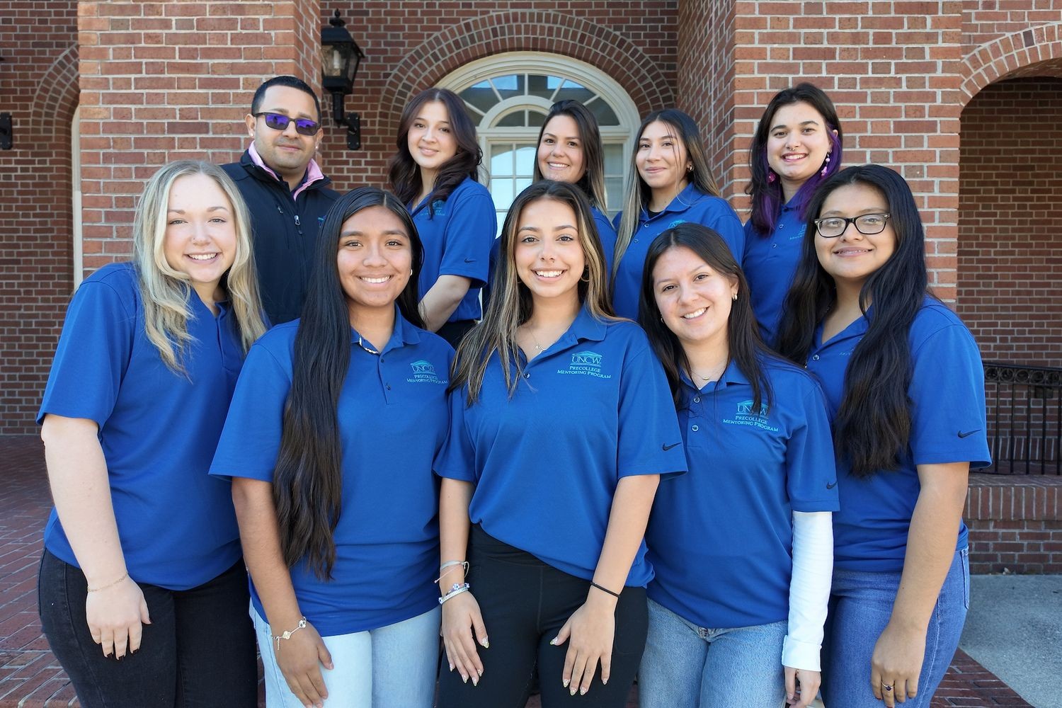 Eight students in blue shirts standing with the director Angel Garcia up together for a team picture