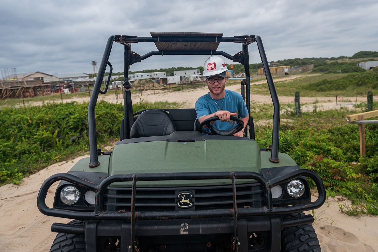 Driver seated in a green utility vehicle on sand, with dunes and buildings in the background
