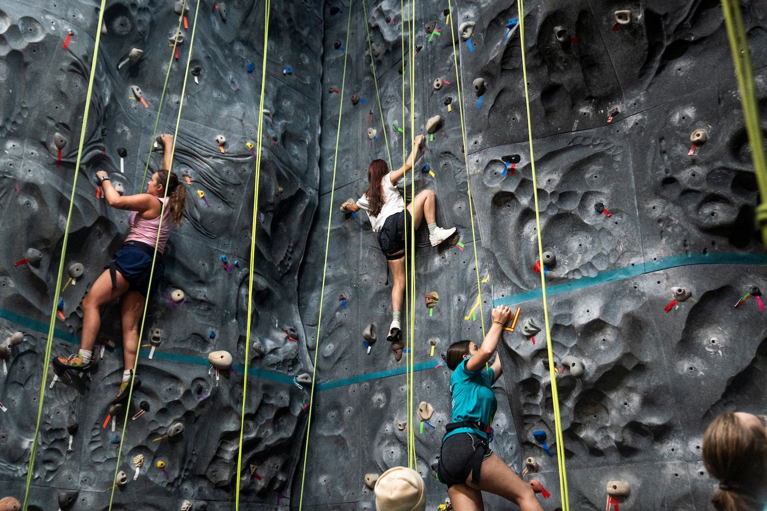 Three people in harnesses climb an indoor rock wall with colorful holds and hanging belay ropes.