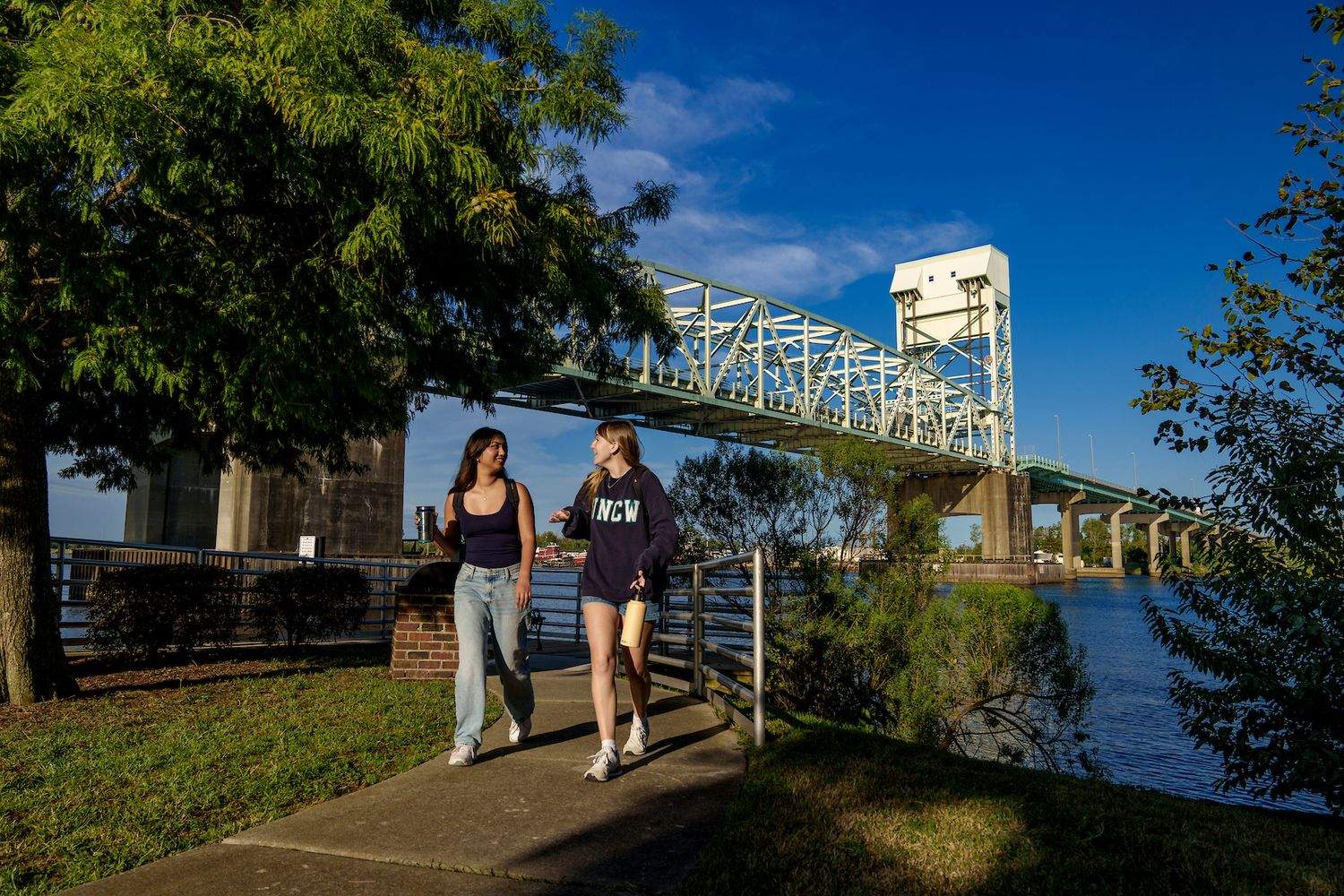 Two people walk along a riverside path with a steel truss bridge and blue sky in the background.