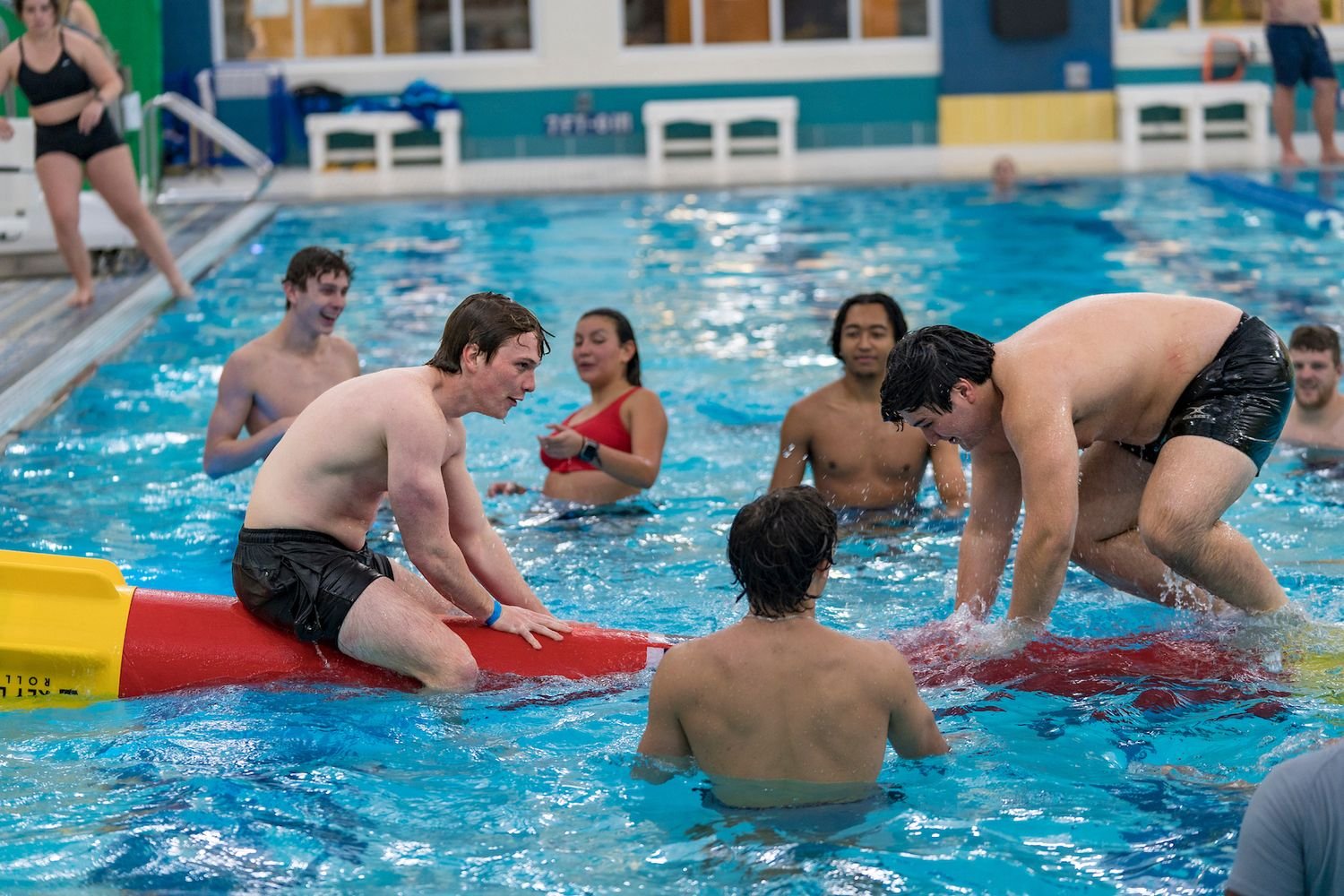 Indoor pool scene with a group playing on a red-and-yellow inflatable log, splashing water.