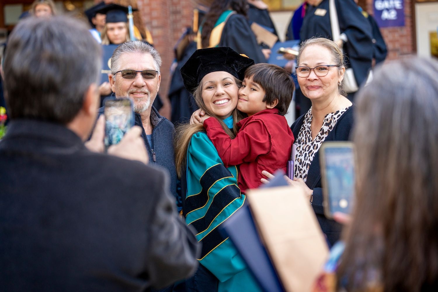 A family takes a photo of a UNCW doctoral graduate in cap and gown, posing with a smiling child in their arms and smiling family members.