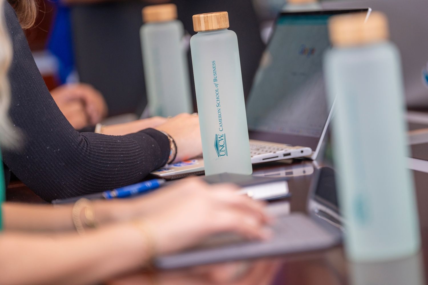 Students typing on laptops with frosted glass UNCW Cameron School of Business water bottles beside them.