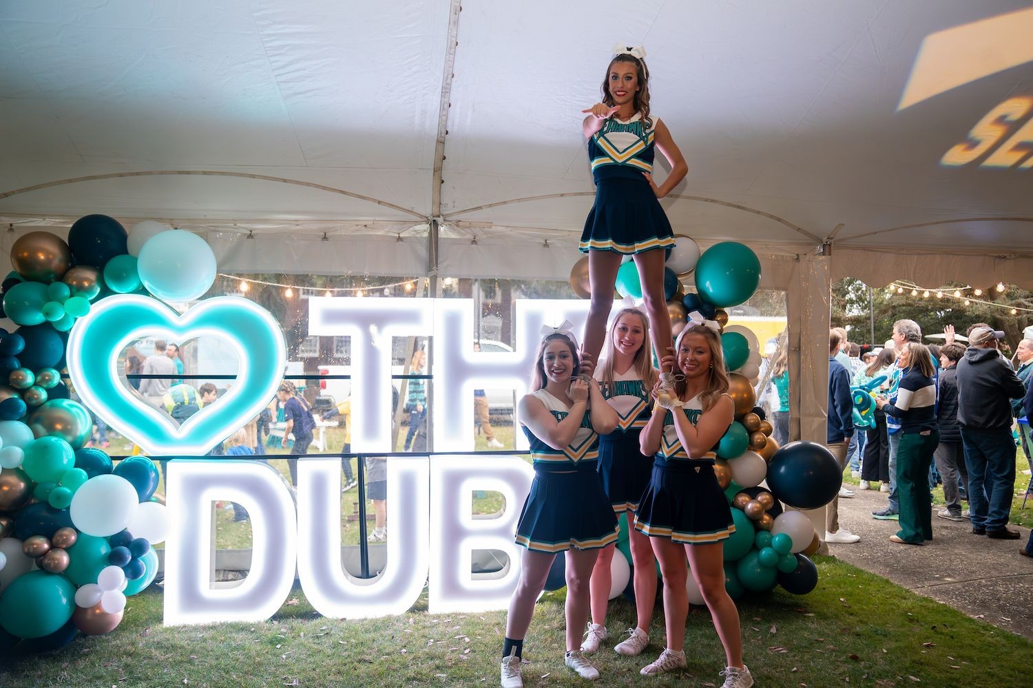 UNCW cheerleaders in a stunt formation beside illuminated "THE DUB" and teal balloons at an outdoor event.