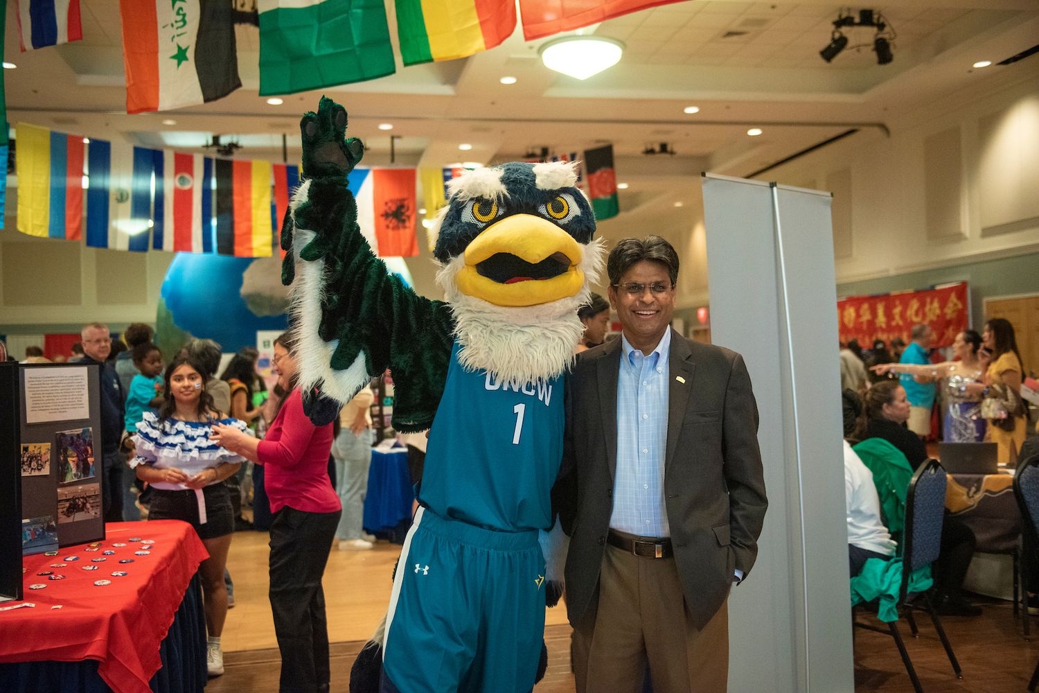 UNCW's Sammy the Seahawk mascot poses with the university's chancellor, amidst a cultural event with international flags displayed overhead.