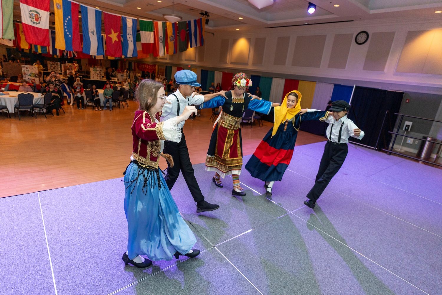 Five children are dancing in traditional ethnic clothing on a stage beneath international flags, likely at a UNC Wilmington event.