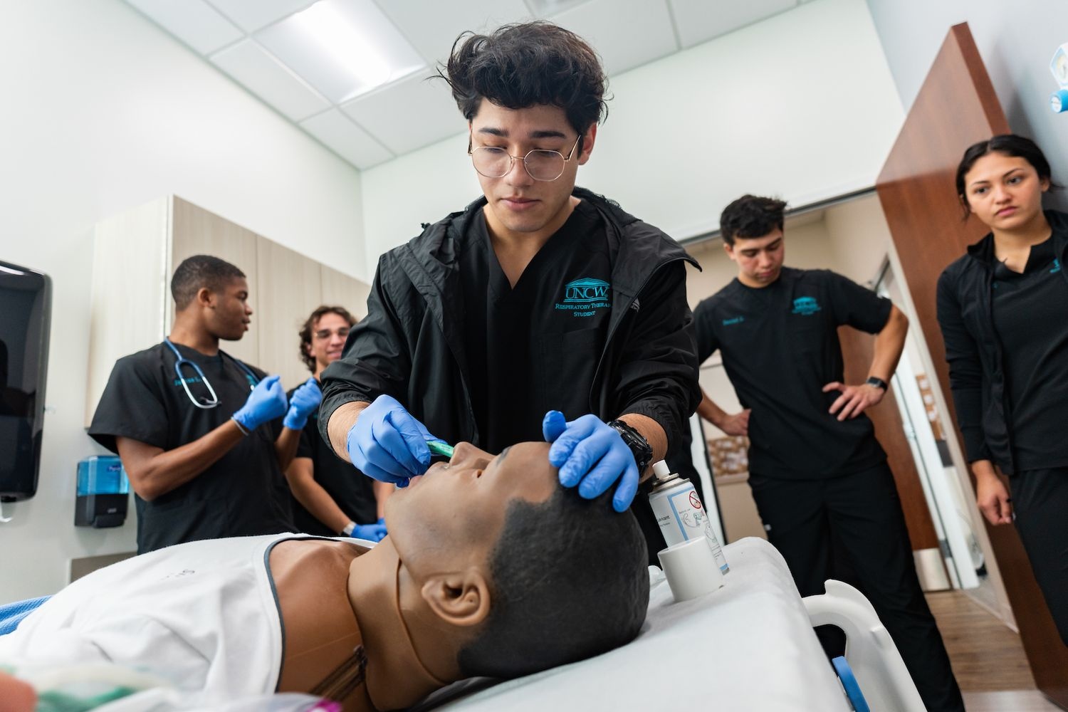 A respiratory therapy student practices intubation on a manikin while fellow respiratory therapy students watch.