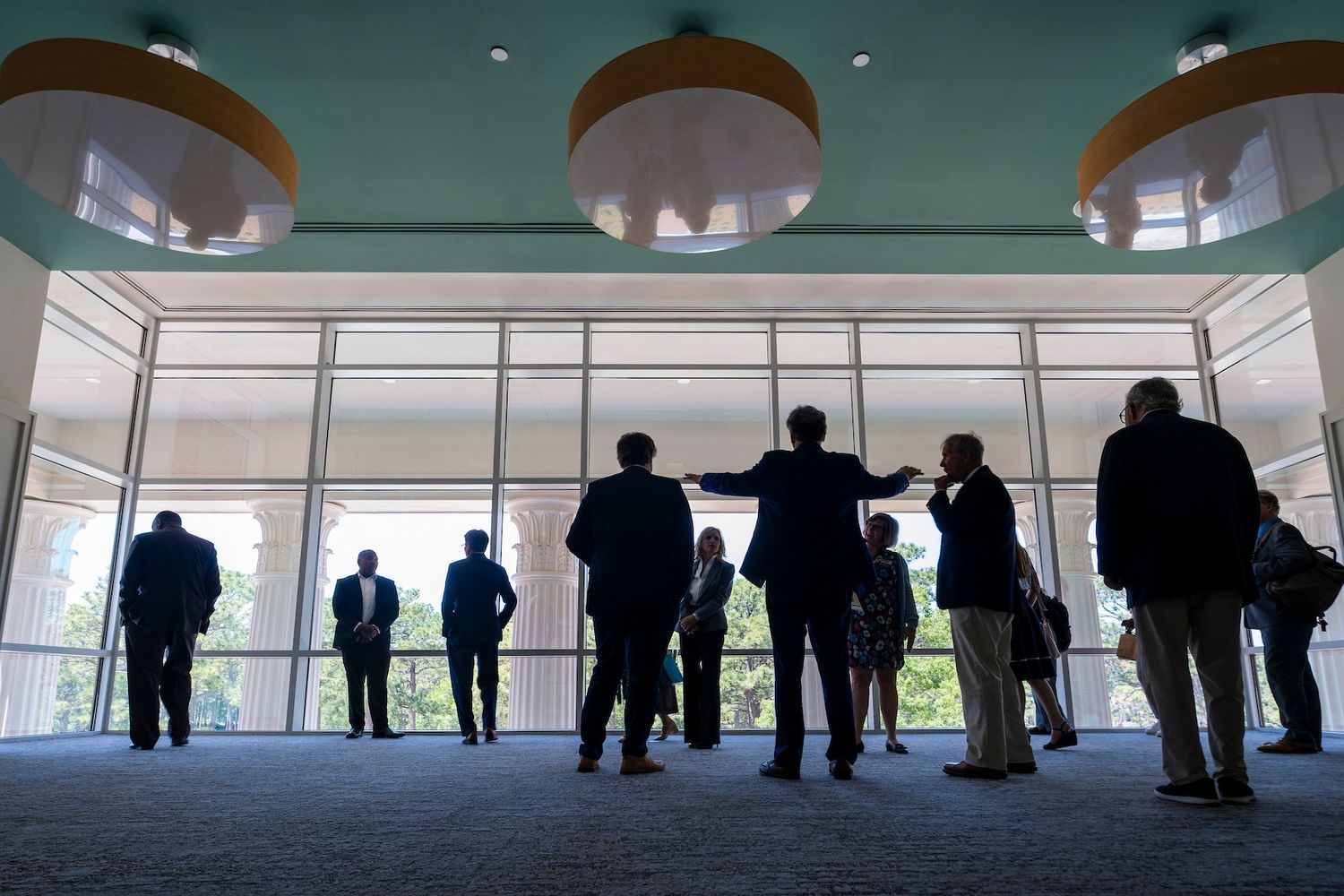 Silhouetted people in business attire stand and talk in a spacious lobby with floor‑to‑ceiling windows and large round ceiling lights.