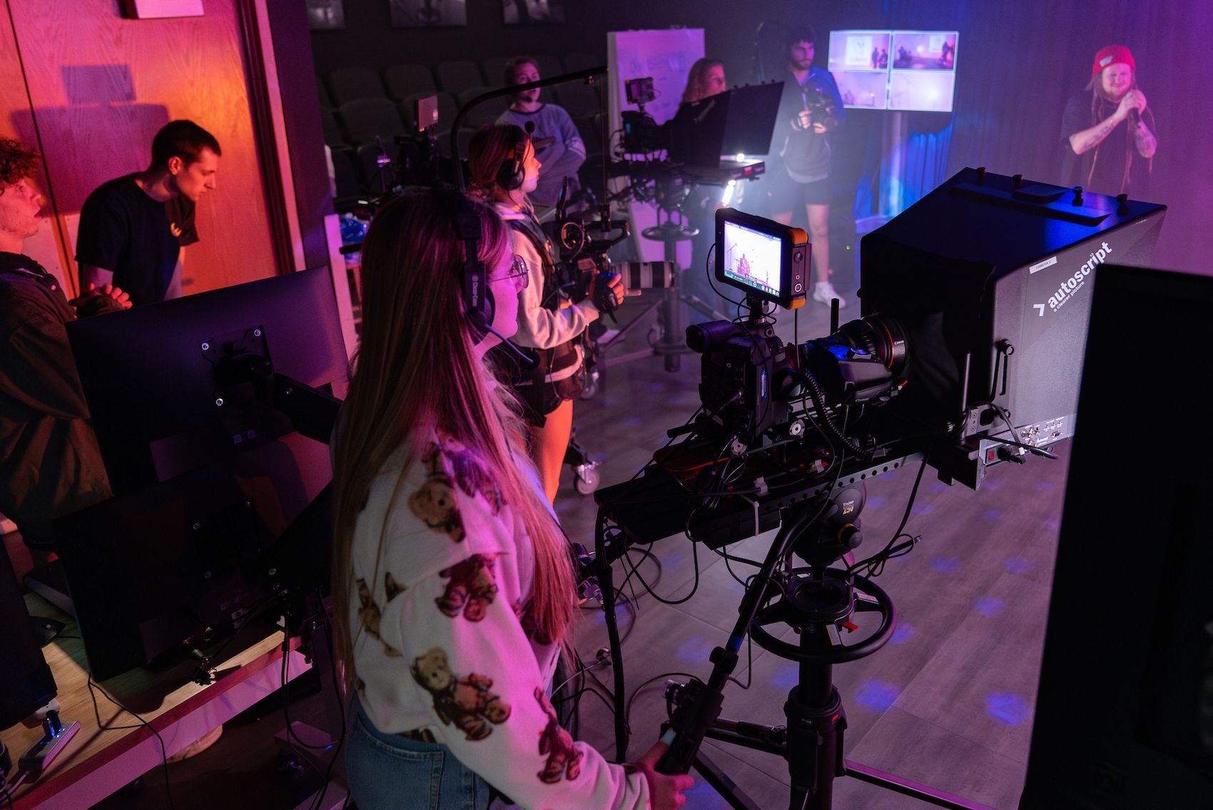 Several students operating tv cameras in a film studio while a musician performs holding a microphone on a stage.