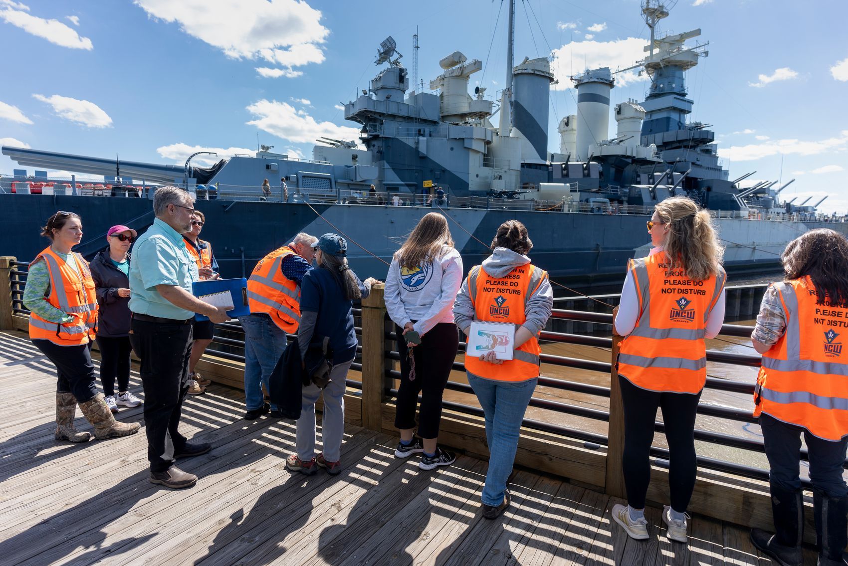 UNCW researchers and students line the railing of the walkway facing Battleship North Carolina.