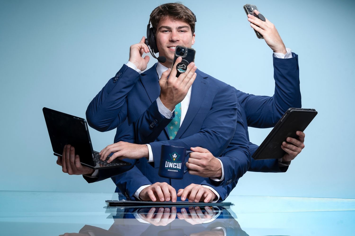 Photo illustration of a young man in a suit with many arms smiling while using cellphones, headset, laptop, tablet and keyboard simultaneously.