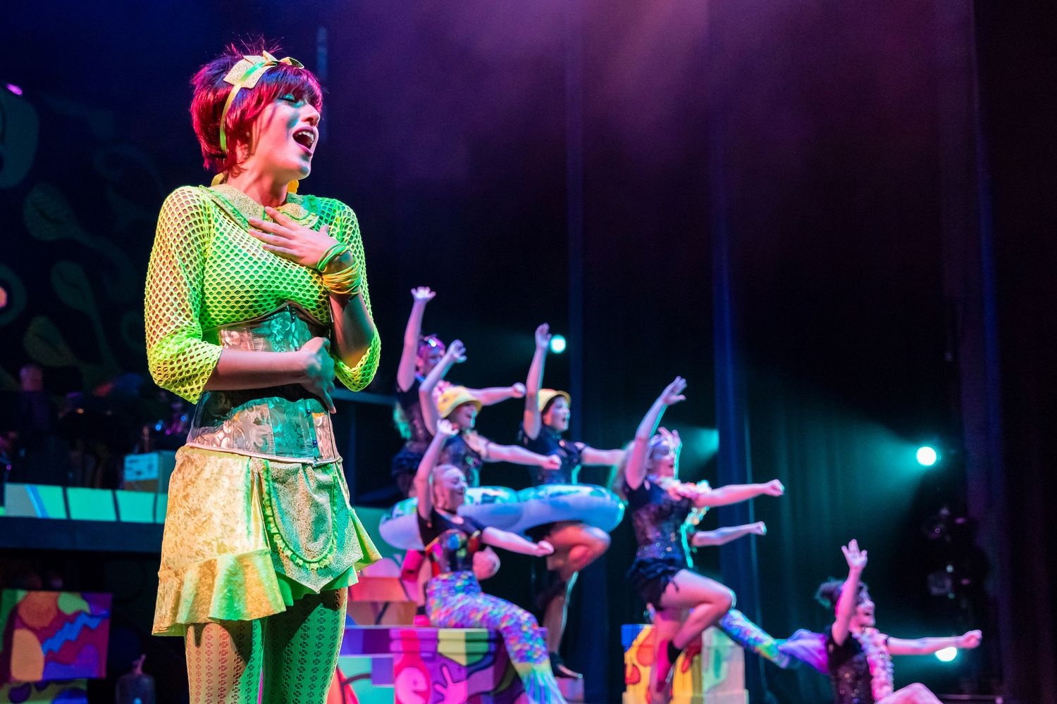 Performer sings on stage during “Head Over Heels” performance with other actors in background.