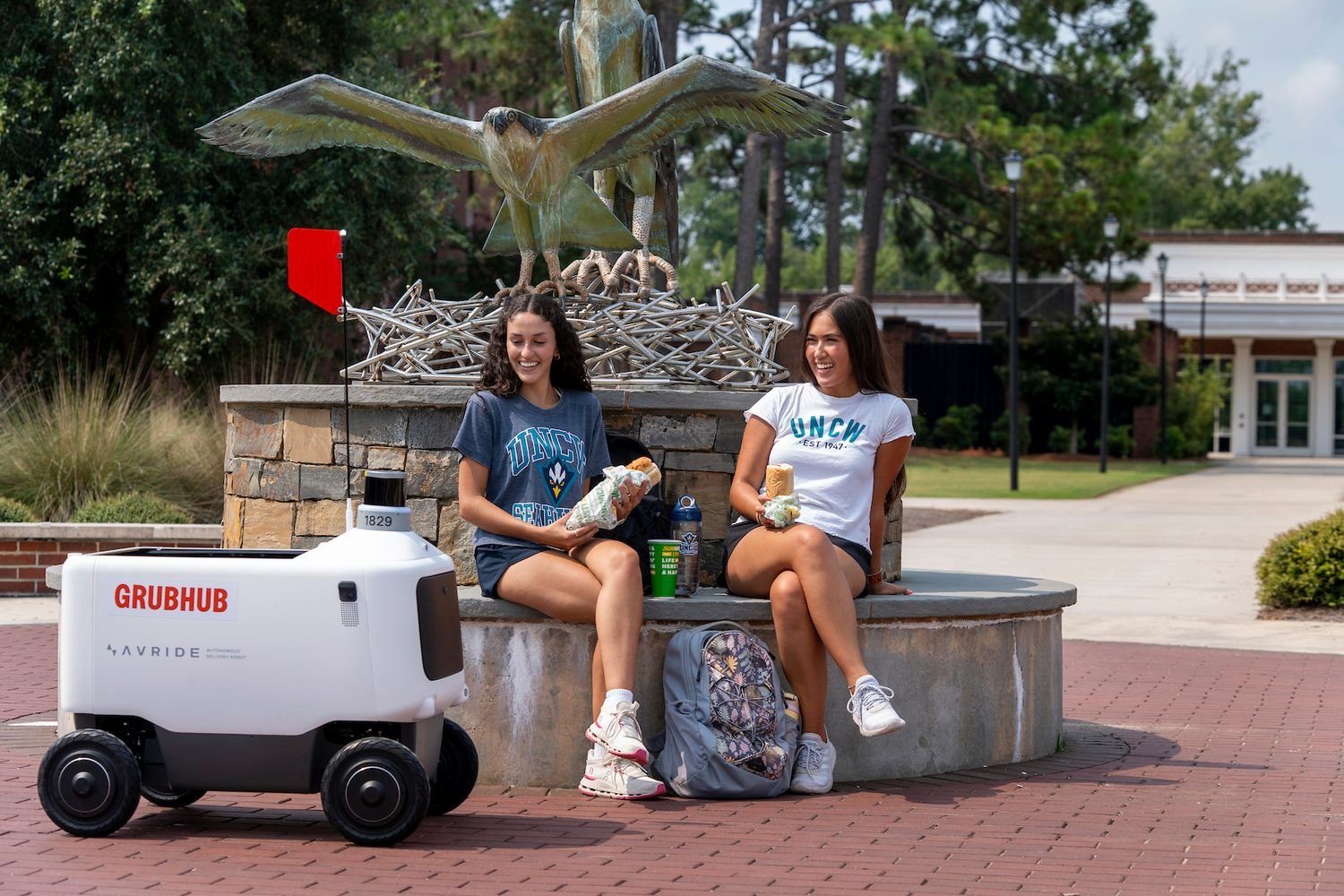 Students sitting  near a statute and a delivery robot on the UNCW campus 