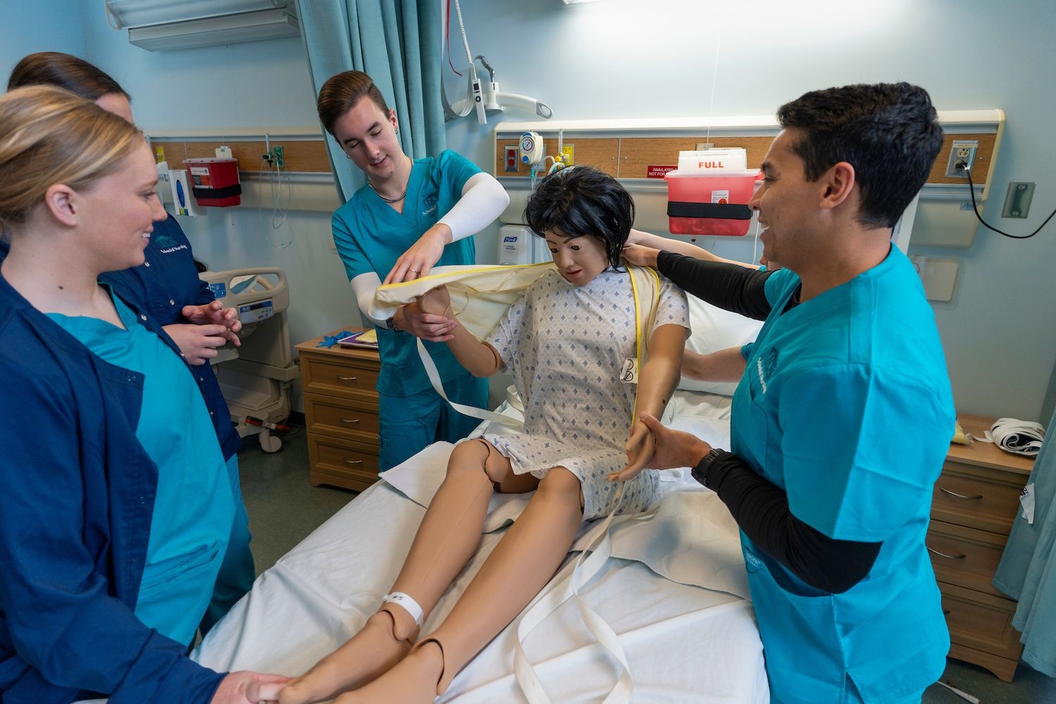 Two nursing students dressed in teal scrubs work together to dress a manikin that's on a hospital bed, while two other students observe.