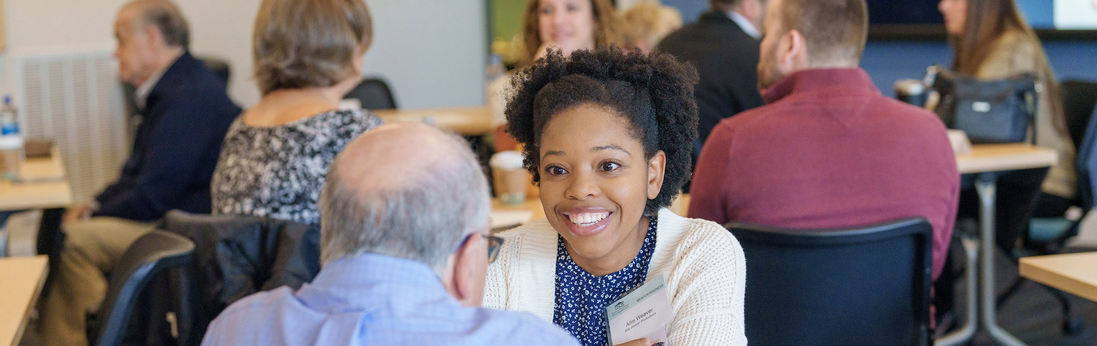 Entrepreneur sitting at a table during an event and engaged in conversation with a mentor seated across from her.