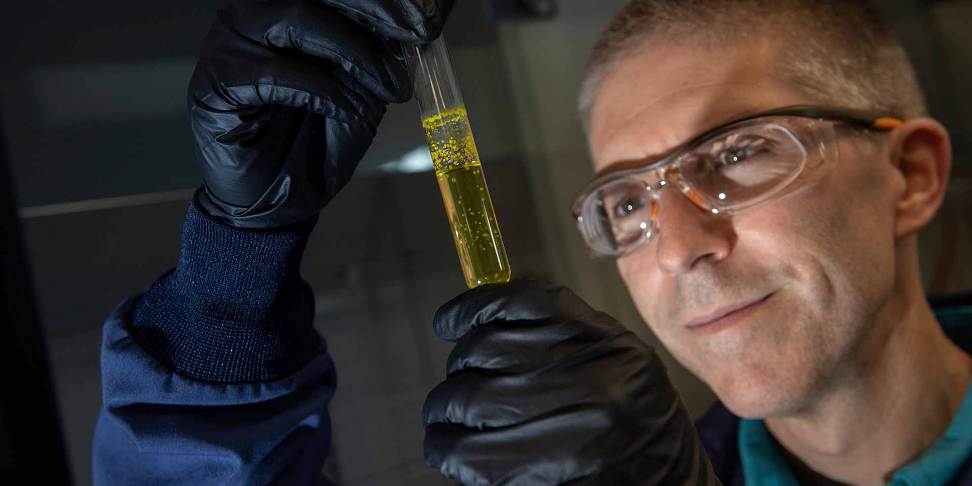 A professor in goggles smiles as he examines a tube of liquid