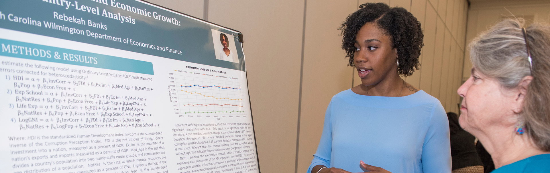 A student standing explains her economic growth research to an onlooker at a poster presentation.