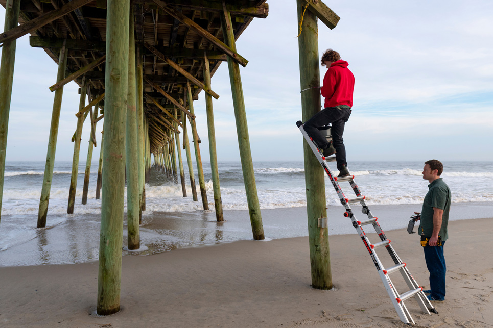 A student standing on a ladder under a pier as another student stands nearby holding a drill.